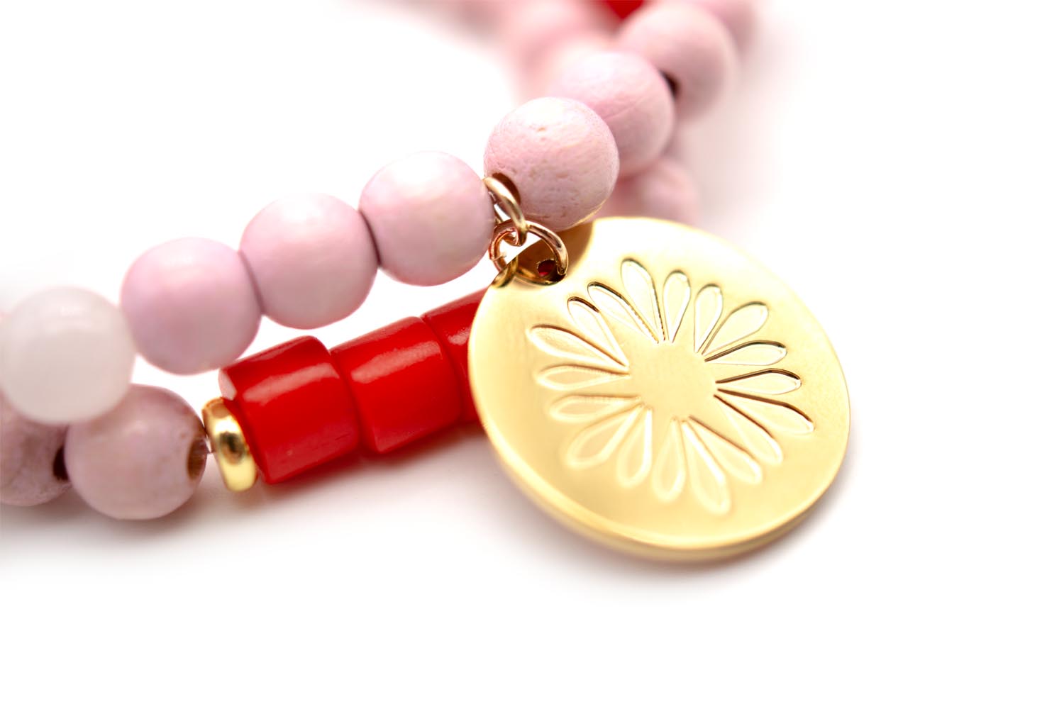 Detailansicht des vergoldeten Logo-Tags mit Blütenmuster neben roten und rosa Perlen