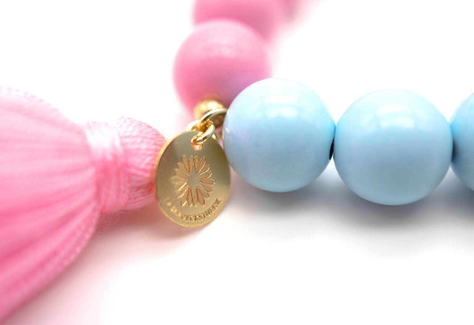 Nahaufnahme der rosa Quaste und der runden goldfarbenen Anhängerplakette neben hellblauen Perlen