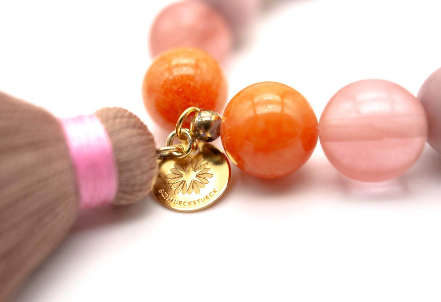 Nahaufnahme der orangefarbenen Jade- und Quarzperlen mit vergoldetem Markenplättchen und rosa umwickelter Quaste