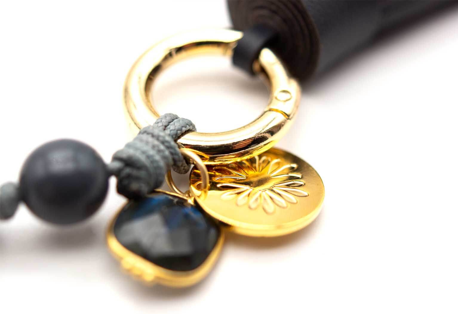 Detail von goldfarbenem Ring, Logo-Münze und Labradorit-Anhänger neben der schwarzen Quaste